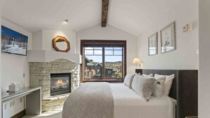 A bedroom with a stone fireplace, a large bed, a desk, and a view of a house through a window.