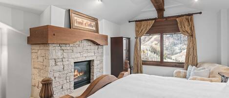 A bedroom with a stone fireplace, a large bed, a wooden headboard, and a view of a snowy landscape through a window.