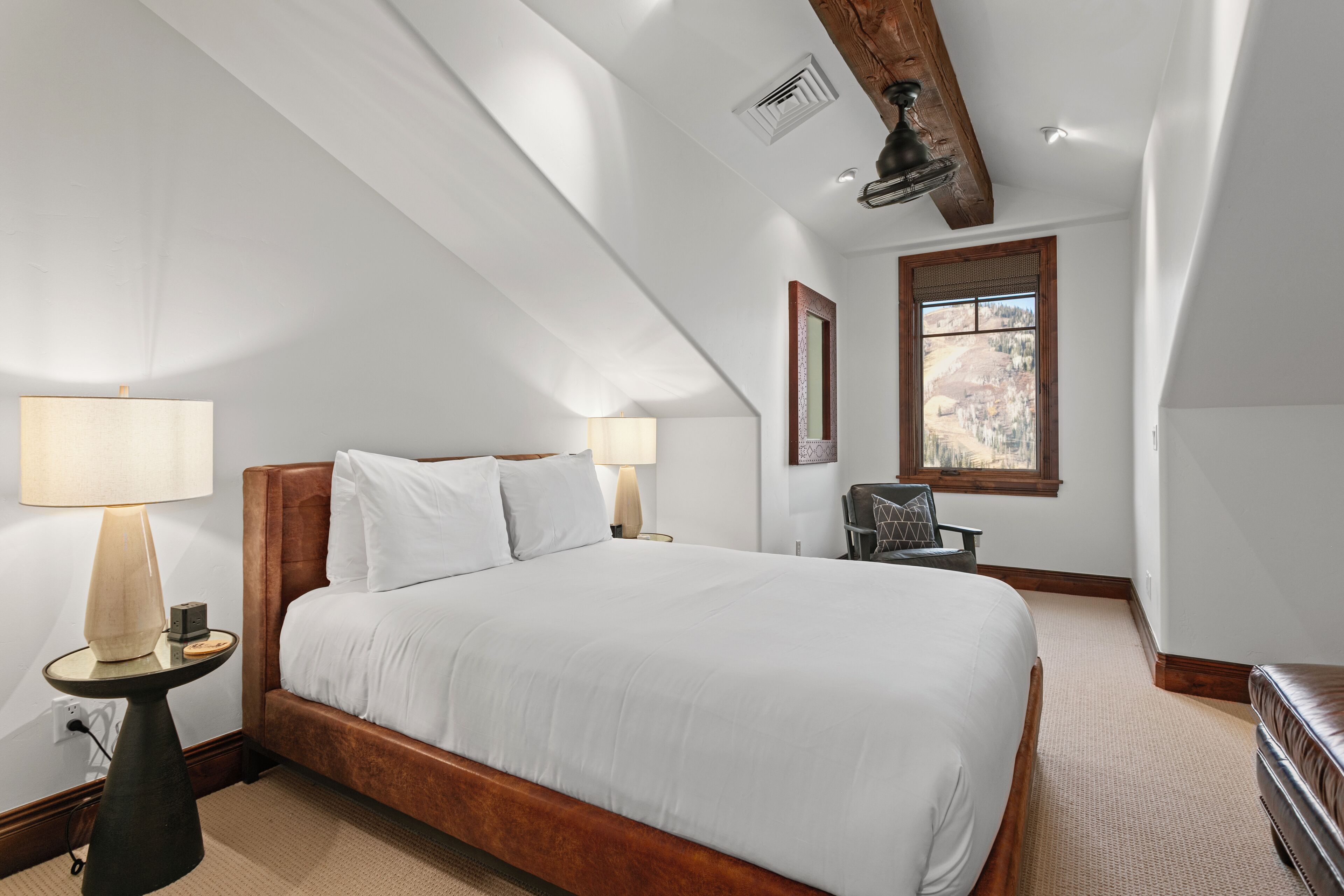 A bedroom with a large bed, two bedside lamps, a window with a view, and a wooden ceiling beam.