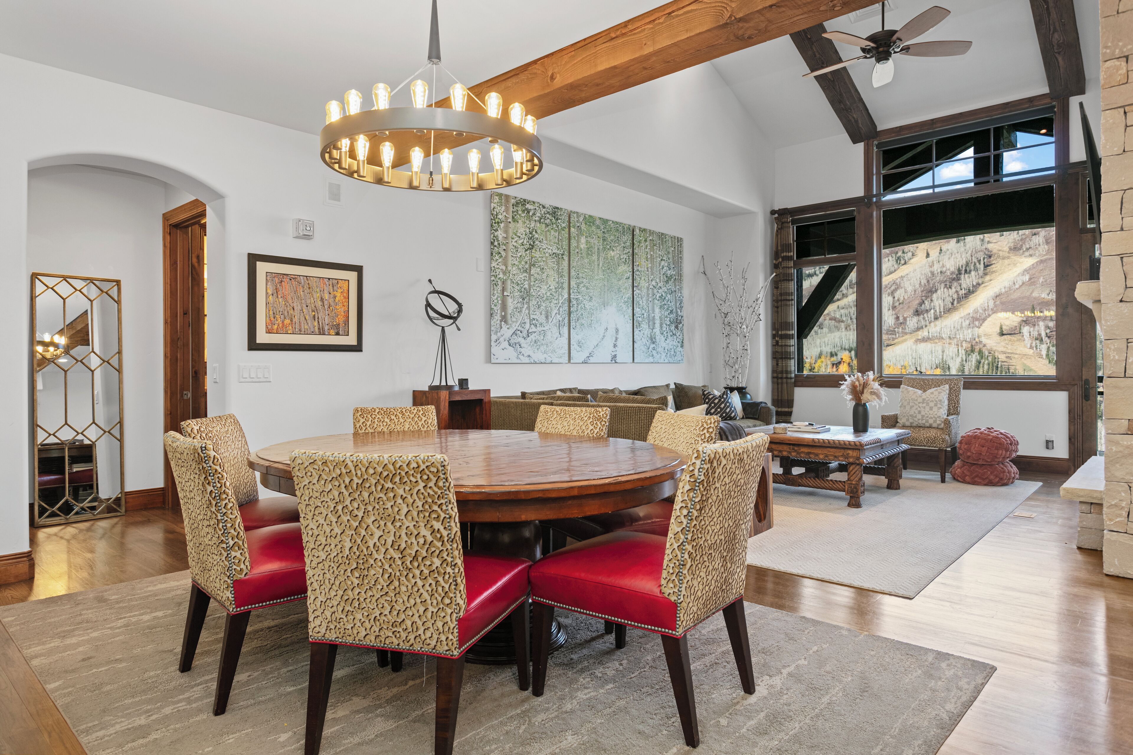 A dining area with a round table and chairs, a chandelier, and a view of a mountainous landscape through large windows.