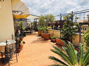 Una terraza en la azotea con baldosas de terracota, plantas en macetas, mobiliario de exterior y un gran paraguas que da sombra.