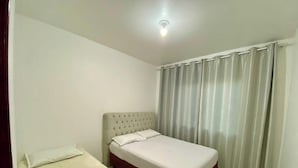 A small, single-bed room with a tufted headboard, a plain white mattress, and a window with sheer curtains. - Casa Aconchego Barra Velha - SC (Barra Velha)