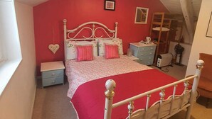Un dormitorio con pared roja, cama blanca con colcha floral, mesita de noche pequeña y mesilla.