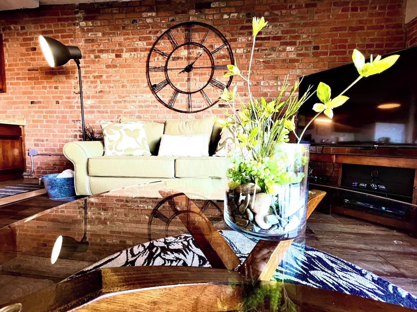 A living room with a brick wall, a sofa, a coffee table with a vase of flowers, a clock, and a flat-screen TV.