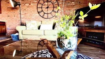 A living room with a brick wall, a sofa, a coffee table with a vase of flowers, a clock, and a flat-screen TV.