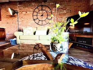 A living room with a brick wall, a sofa, a coffee table with a vase of flowers, a clock, and a flat-screen TV. - Waterfront W/ Patio Amazing View Cedar Point (Sandusky)