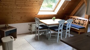 Una habitación acogedora con techo de madera, tragaluces, una mesa con sillas, un radiador y un cubo de basura pequeño.