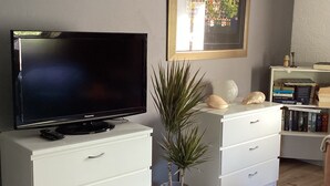 Una sala de estar con una televisión de pantalla plana sobre un mueble blanco, una planta en maceta, una estantería con libros y un cuadro en la pared.