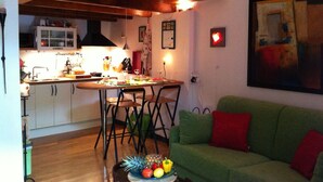 Un coin repas avec une table en bois, des chaises et un canapé vert. On aperçoit une cuisine en arrière-plan, avec des placards et une cuisinière.
