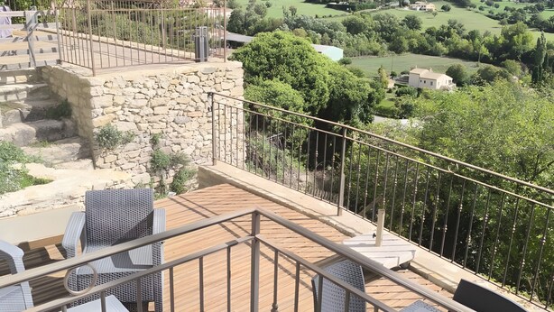 Un balcón con mobiliario exterior, un muro de piedra y vistas a un valle verde y montañas.
