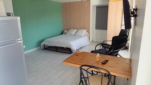 Una habitación pequeña con una cama, una mesa, dos sillas, un refrigerador y una cortina.