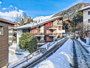 A snow-covered street with buildings, including a multi-story structure with balconies and a wooden building with a sign. - Verte-Rive by Interhome (Zermatt)