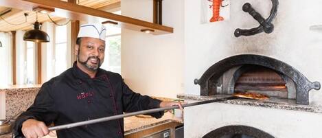 Un chef junto a un horno de pizza de ladrillo, una decoración de langosta y un estante de metal.