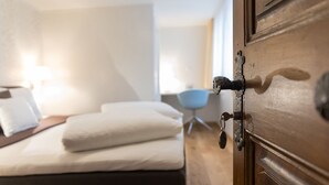 A wooden door with a keyhole and a lock, a bed with white linens, a bedside table, and a lamp in a hotel room.