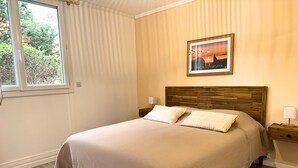 Una cama bien hecha con cabecera de madera, dos mesitas de noche con lámparas, una ventana con persianas y un cuadro enmarcado en la pared.