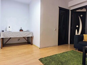 Una habitación con piso de madera, una pequeña mesa con mantel, una puerta negra y un sofá de color oscuro.