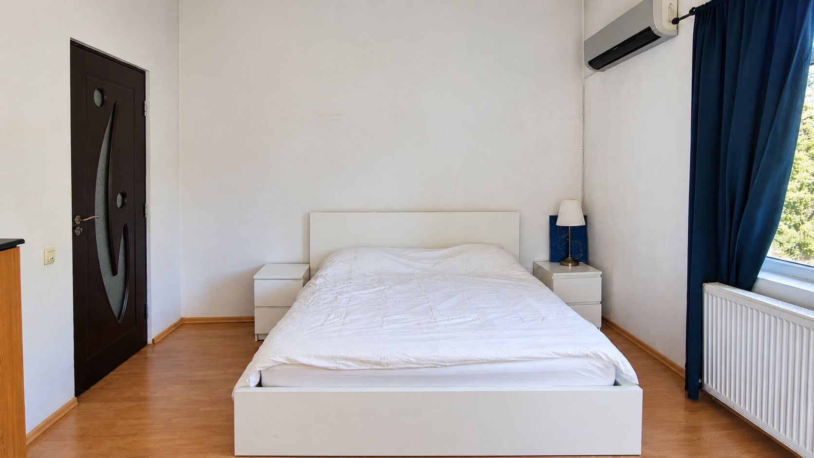 Una camera da letto con un letto singolo, due comodini, un tavolino e una finestra con tende, oltre a un condizionatore d'aria.