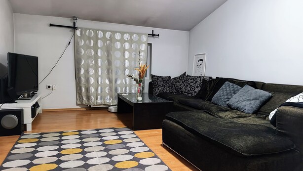 Een moderne woonkamer met een zwarte bank, een bijzettafeltje, een tv op een meubelstuk, een kamerbreed tapijt en een gordijn met ronde uitsparingen.