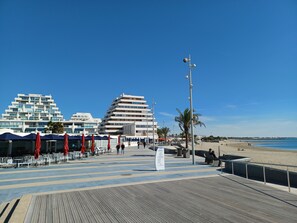 Eine Küstenpromenade mit modernen Gebäuden, Außenbestuhlung und einem Strand im Hintergrund.