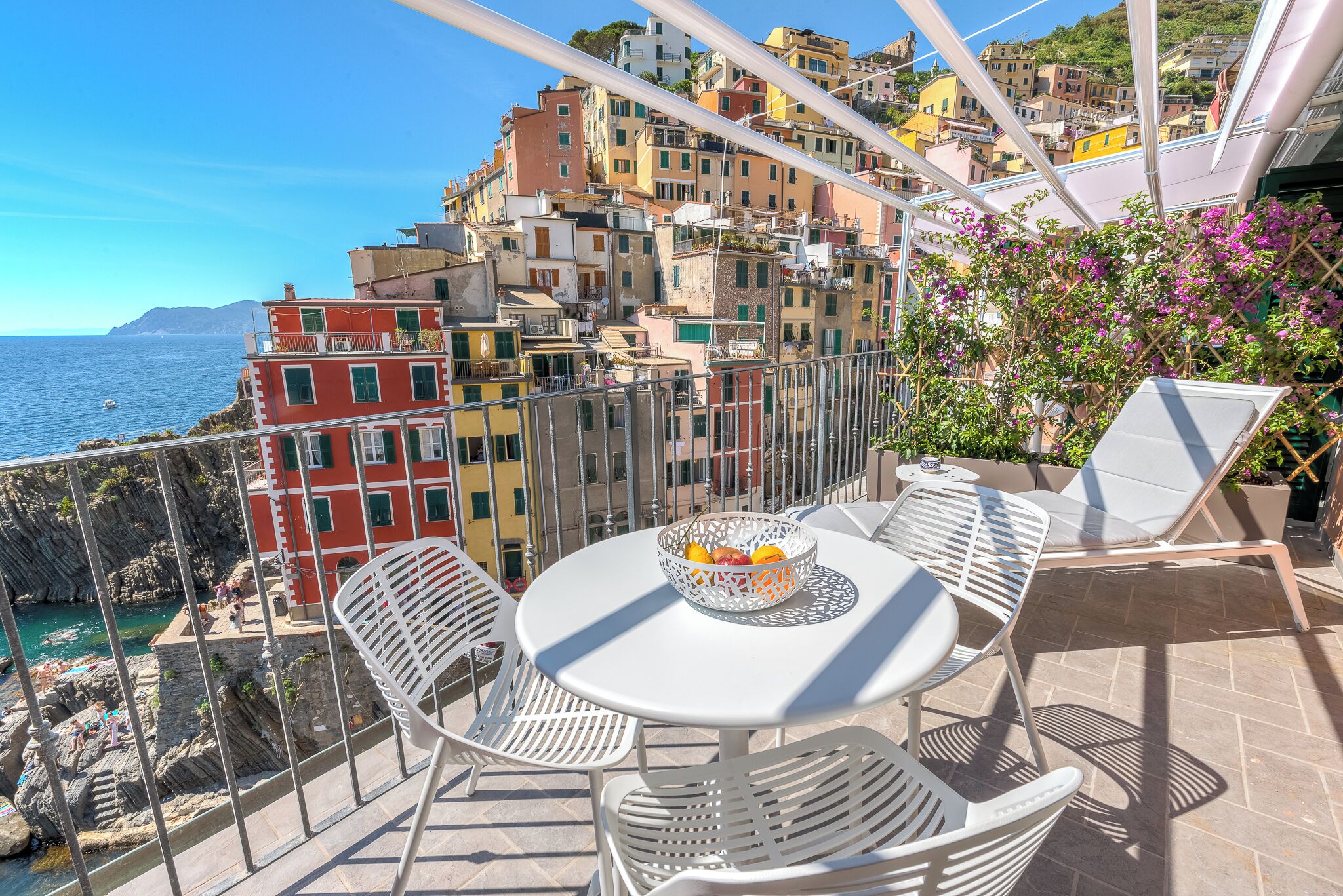 Une terrasse avec une table et des chaises, d’où l’on peut admirer une ville côtière aux bâtiments aux couleurs vives et la mer.