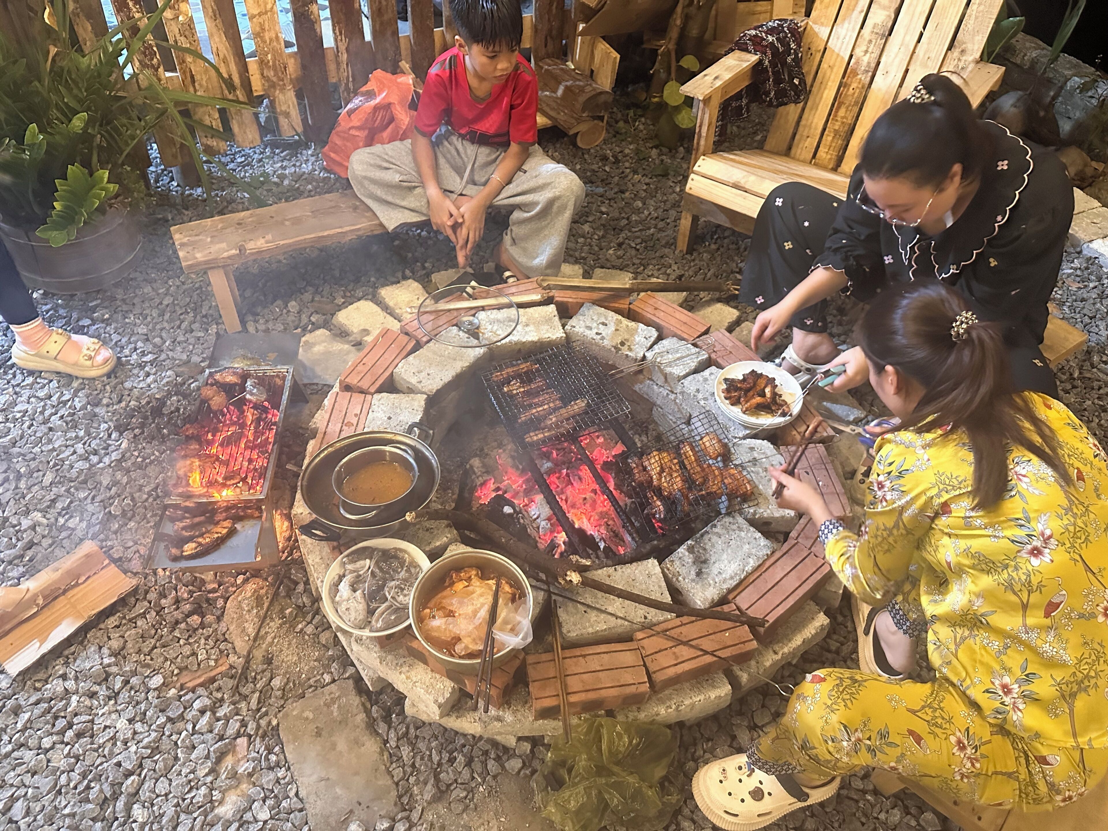 A circular fire pit with food being cooked, surrounded by people.