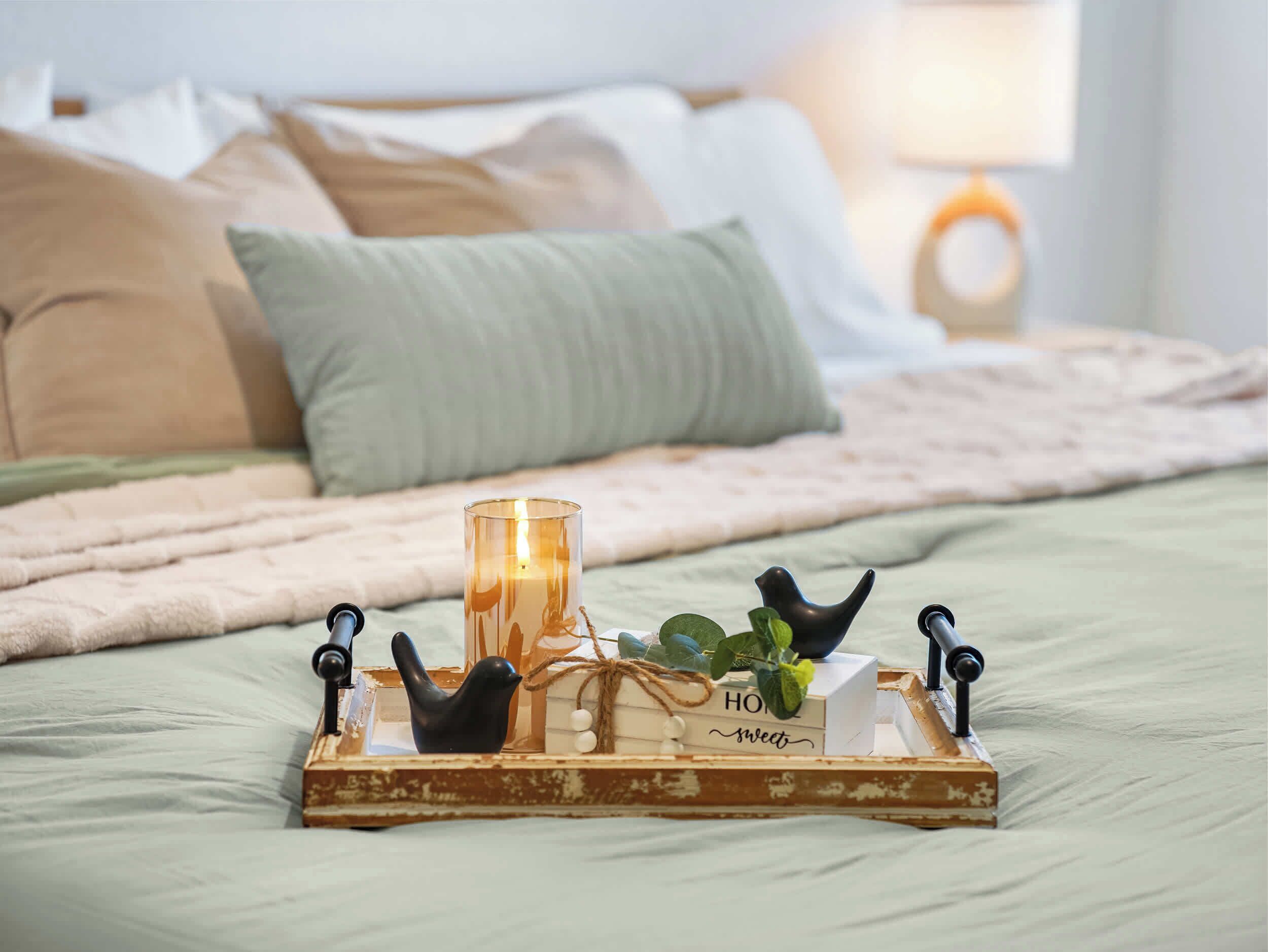 A bed with a wooden tray holding a candle, a bird figurine, and a small plant.