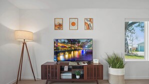 Een woonkamer met een televisie op een houten standaard, een vloerlamp en drie kunstwerken in lijsten aan de muur.