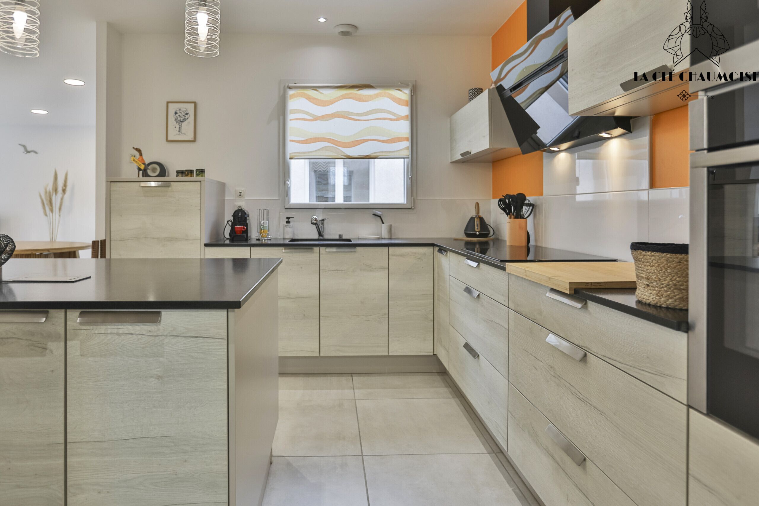 A modern kitchen with a central island, stainless steel appliances, and a window with a wavy pattern blind.