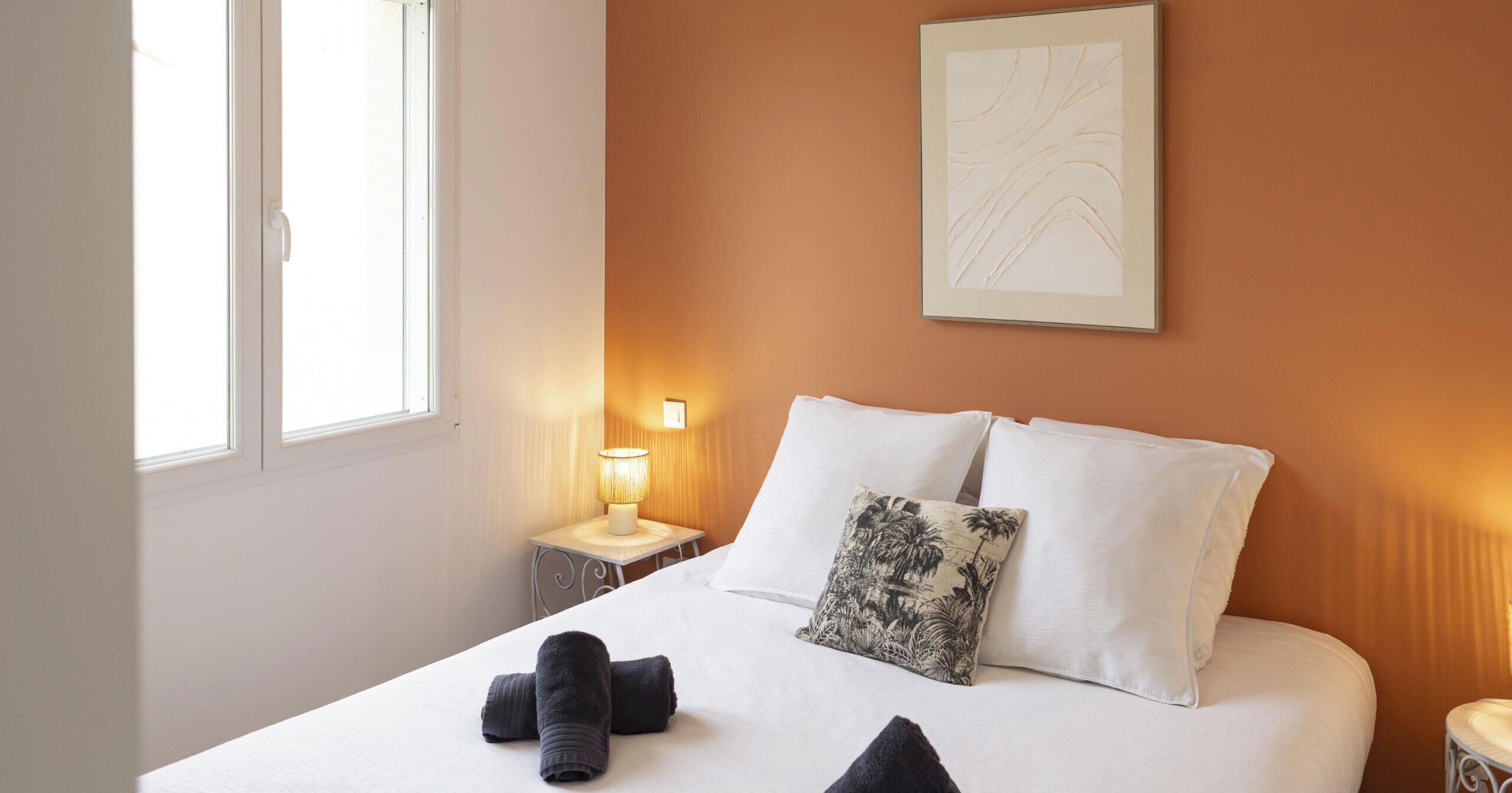 Un lit bien fait, avec du linge de lit blanc et deux oreillers, dont l’un est orné d’un motif tropical. Une lampe se trouve sur une table de chevet à gauche, et une œuvre d’art encadrée est accrochée au mur au-dessus du lit.