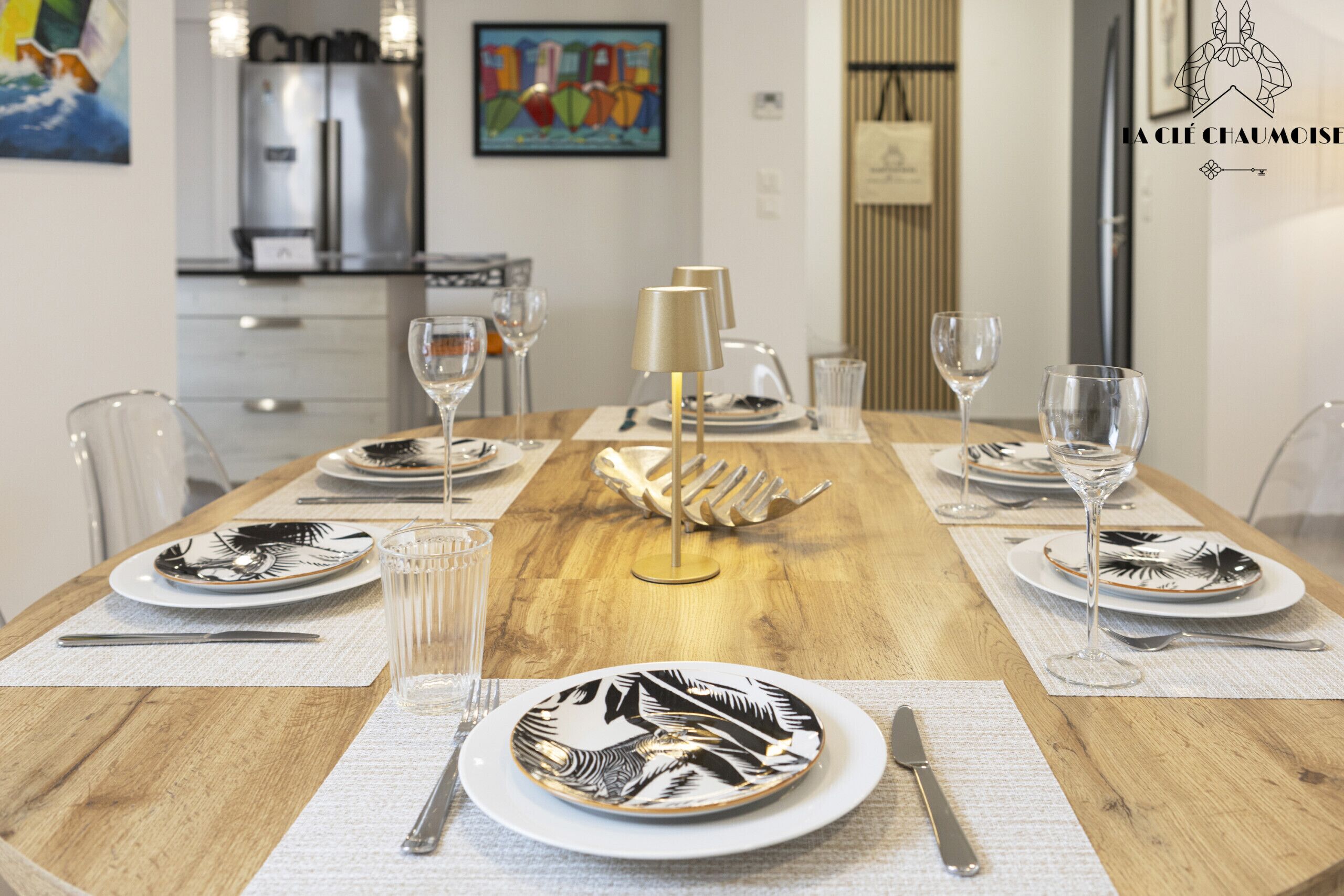 Une table à manger dressée avec des assiettes, des couverts et des verres, avec une cuisine moderne en arrière-plan.