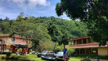Um cenário tranquilo às margens do rio, com um barco pequeno atracado perto de um edifício, uma pessoa parada à beira da água e uma paisagem verde exuberante ao fundo.