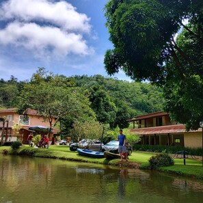 A serene riverside setting with a small boat docked near a building, a person standing by the water, and a lush green landscape in the background. - Pousada Pedra d'água (Guarapari)