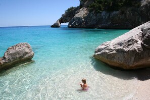 A beach with clear turquoise water, large rocks, and a child playing in the shallow water.