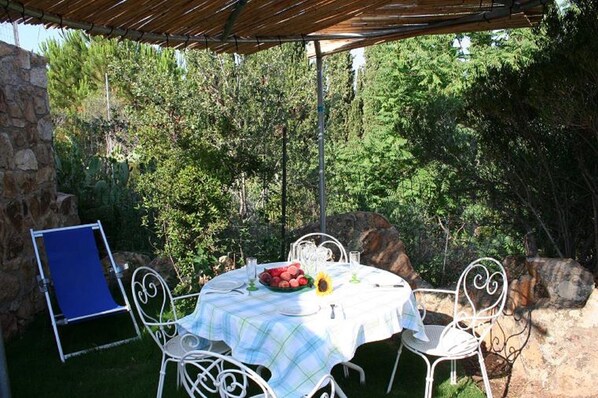 An outdoor dining area with a table set for two, surrounded by white metal chairs, under a wooden pergola.