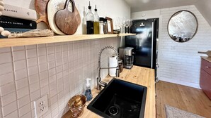 A modern kitchen with a wooden countertop, a black sink, and a stainless steel refrigerator. There are shelves above the countertop holding various kitchen items and a mirror on the wall. - Serene 2-bedroom A-Frame in Jefferson perfect for Hot Tub + Views + Stargazing (Jefferson)