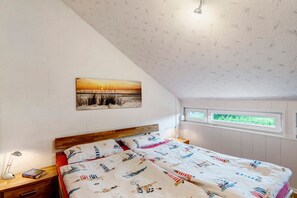 Et soveværelse med skrå væg, en træsenggavl, en seng med et quiltet betræk i maritimt tema, et natbord med en lampe og et vindue med udsigt over grøn bevoksning.