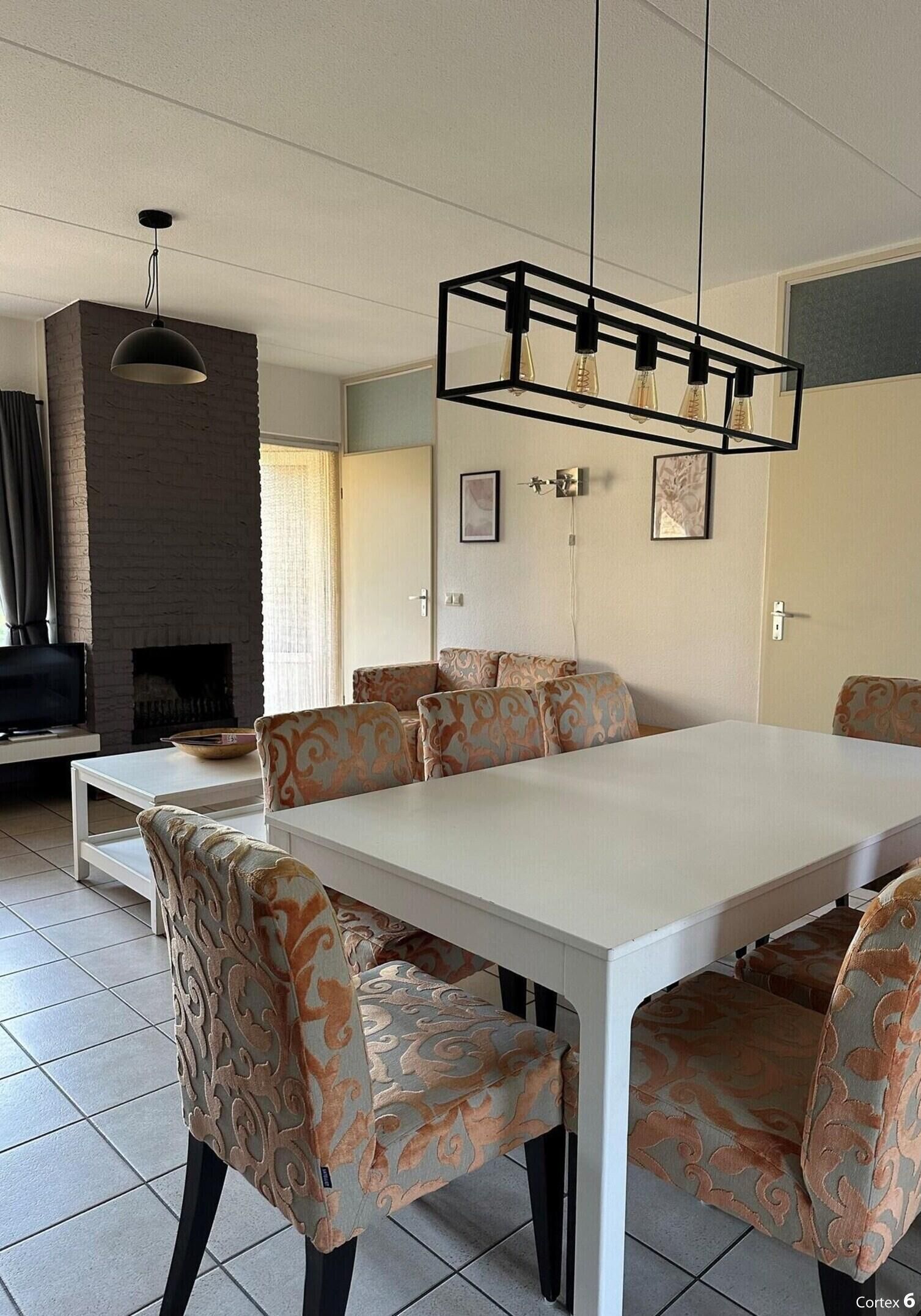 Un comedor con una mesa blanca y sillas estampadas, una chimenea de ladrillo y una lámpara colgante.