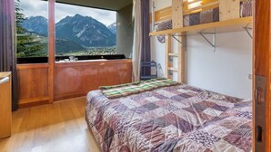 Une chambre avec une grande fenêtre, un lit recouvert d’une courtepointe, un lit superposé en bois et une vue sur les montagnes.