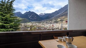 Un balcon donnant sur la ville et les montagnes, une table en bois avec des tasses et une cafetière, et une chaise.