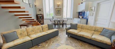 Sala de estar com dois sofás, uma mesa de centro e uma escada. Há também uma área de jantar com mesa e cadeiras ao fundo.