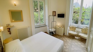 Habitación de hotel con cama, radiador, escritorio, silla, televisor y ventanas amplias que dan a zonas verdes y montañas.