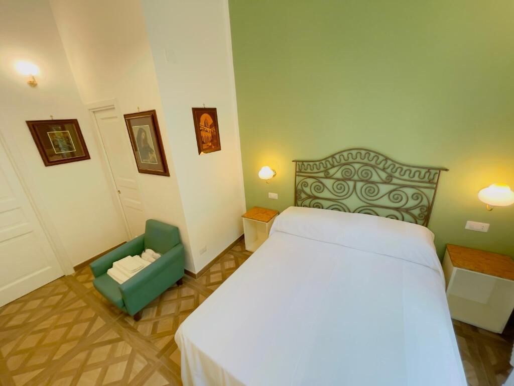 A hotel room with a bed, a green armchair, two bedside tables, and wall-mounted lamps.