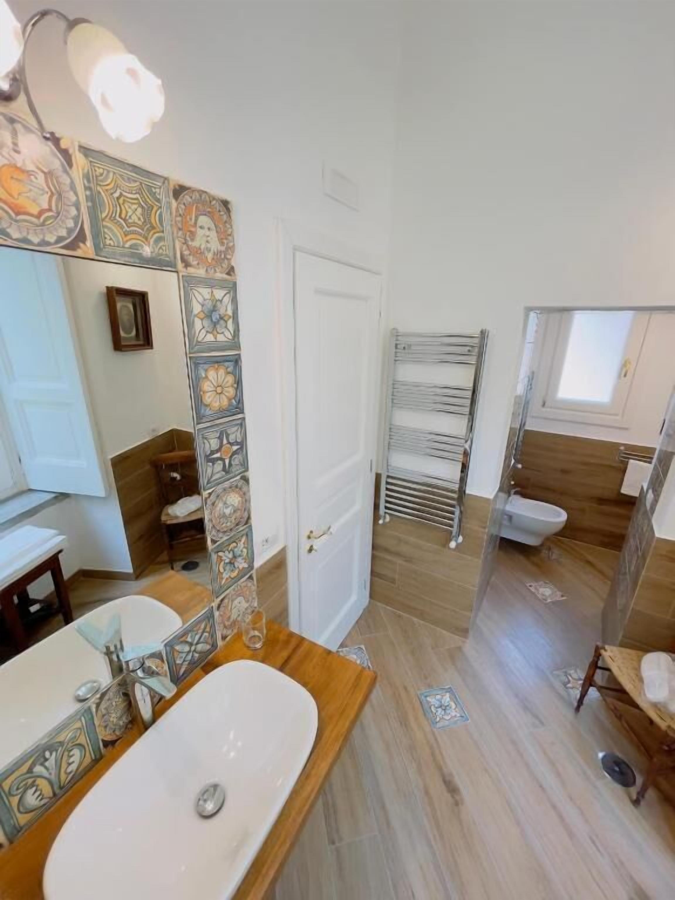 Een badkamer met een houten wastafelmeubel, een spiegel met decoratieve tegels, een witte deur en een douchecel met een glazen deur.