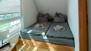 Um quarto pequeno e inclinado com uma cama, travesseiros, uma escrivaninha com um computador e uma janela com persianas.
