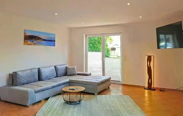 A living room with a grey sectional sofa, a round coffee table, a painting on the wall, a floor lamp, and a sliding glass door leading to a green outdoor area. - Holiday Home Rechlin Lake Apartment 3 (Rechlin)