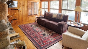 A cozy log cabin interior with a wooden ceiling, a fireplace, a large window, and a comfortable seating area. - The Hidey-Ho on Lower Clam Lake | Secluded | Private Dock (Clam Lake)