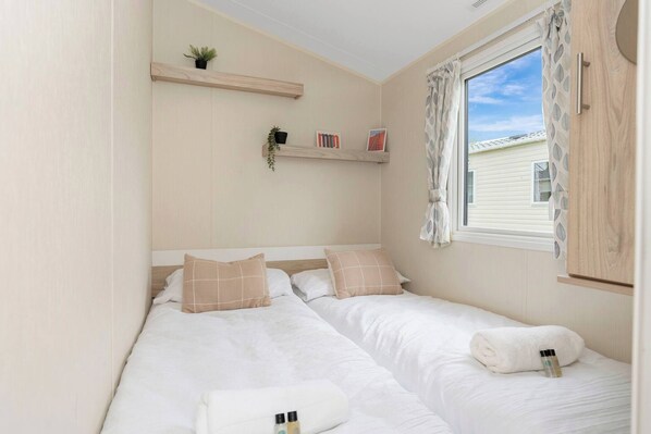 A bedroom with two beds, a window with curtains, a shelf with plants and books, and a towel on one bed.