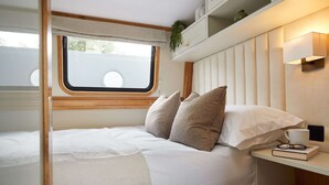 A compact, well-lit bedroom with a bed, pillows, a bedside table with a book and glasses, and a window with a view. - Schwimmendes Wohnboot in Berlin (Berlin)