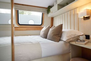A compact, well-lit bedroom with a bed, pillows, a bedside table with a book and glasses, and a window with a view.