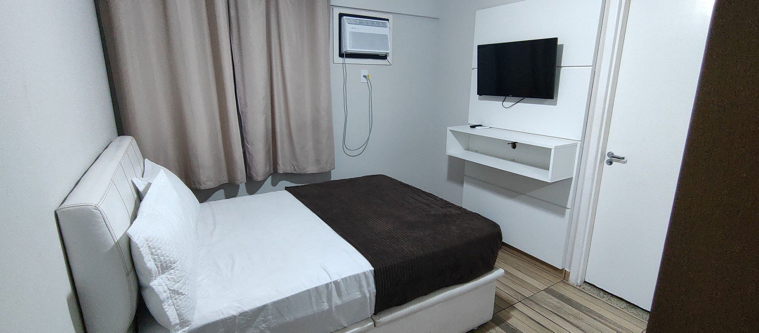 A small, single-bed room with a white headboard, a bedside table, a flat-screen TV mounted on the wall, and a window with curtains.